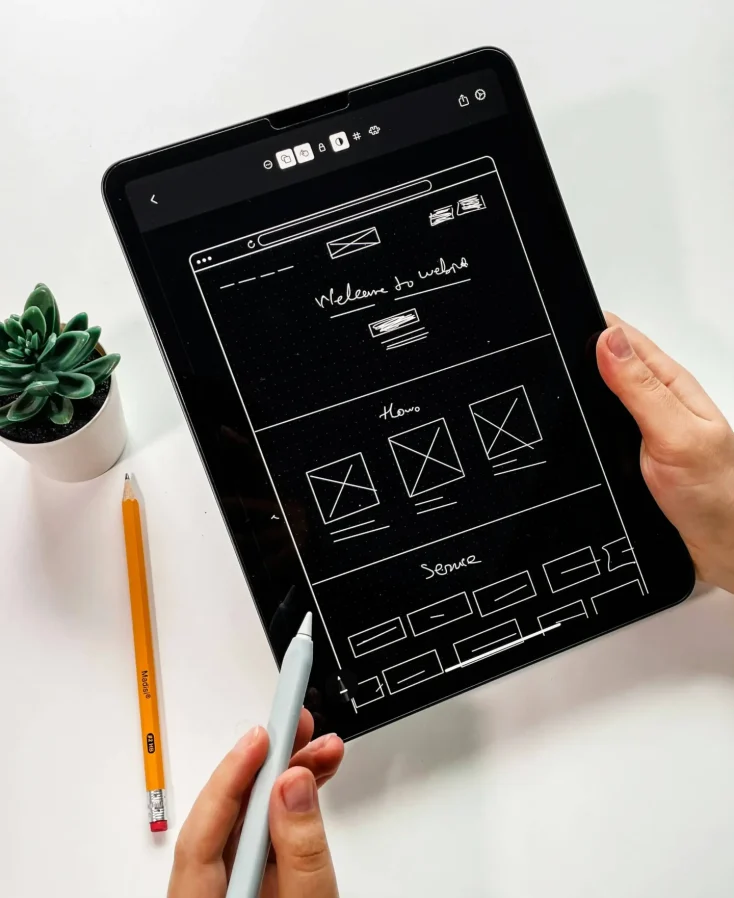 Web Design Hands holding a tablet and a stylus. A plastic plant is nearby.